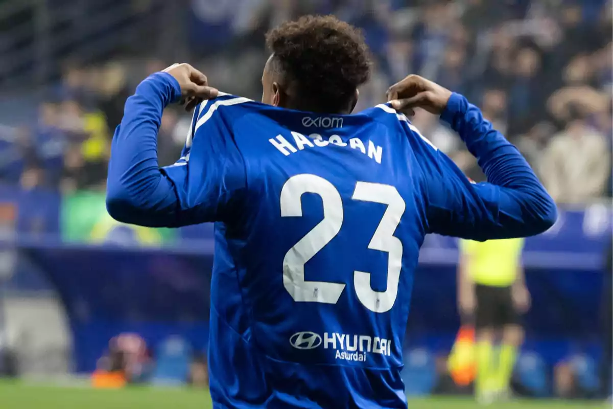 Jugador de fútbol celebrando y mostrando la parte trasera de su camiseta azul con el nombre Hassan y el número 23 en un estadio lleno de espectadores. Jugador de fútbol celebrando y mostrando la parte trasera de su camiseta azul con el nombre Hassan y el número 23 en un estadio lleno de espectadores.