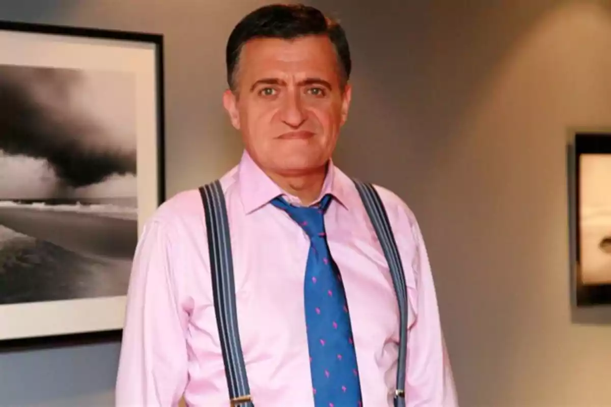 Hombre de cabello corto y oscuro con camisa rosa, tirantes y corbata azul, posando en una habitación con cuadros en la pared Hombre de cabello corto y oscuro con camisa rosa, tirantes y corbata azul, posando en una habitación con cuadros en la pared