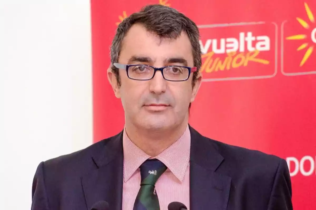 Hombre de cabello corto y gafas oscuras con traje y corbata de pie frente a un fondo rojo con letras blancas y amarillas