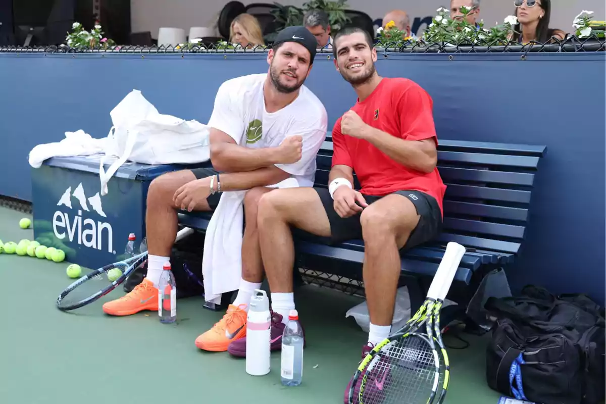 Dos tenistas sentados en una banca sonríen y posan con el brazo flexionado en una cancha de tenis, rodeados de raquetas, botellas de agua y pelotas, mientras varias personas los observan desde el fondo.