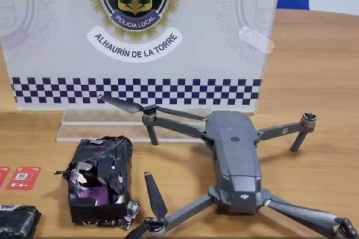 Un dron gris junto a dos paquetes envueltos en cinta negra y dos tarjetas SIM sobre una mesa de madera, con un cartel de la policía local de Alhaurín de la Torre al fondo. Un dron gris junto a dos paquetes envueltos en cinta negra y dos tarjetas SIM sobre una mesa de madera, con un cartel de la policía local de Alhaurín de la Torre al fondo.