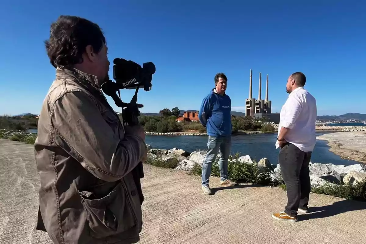 Un camarógrafo graba a dos hombres conversando al aire libre cerca de un río con una central eléctrica de tres chimeneas al fondo en un día soleado. Un camarógrafo graba a dos hombres conversando al aire libre cerca de un río con una central eléctrica de tres chimeneas al fondo en un día soleado.