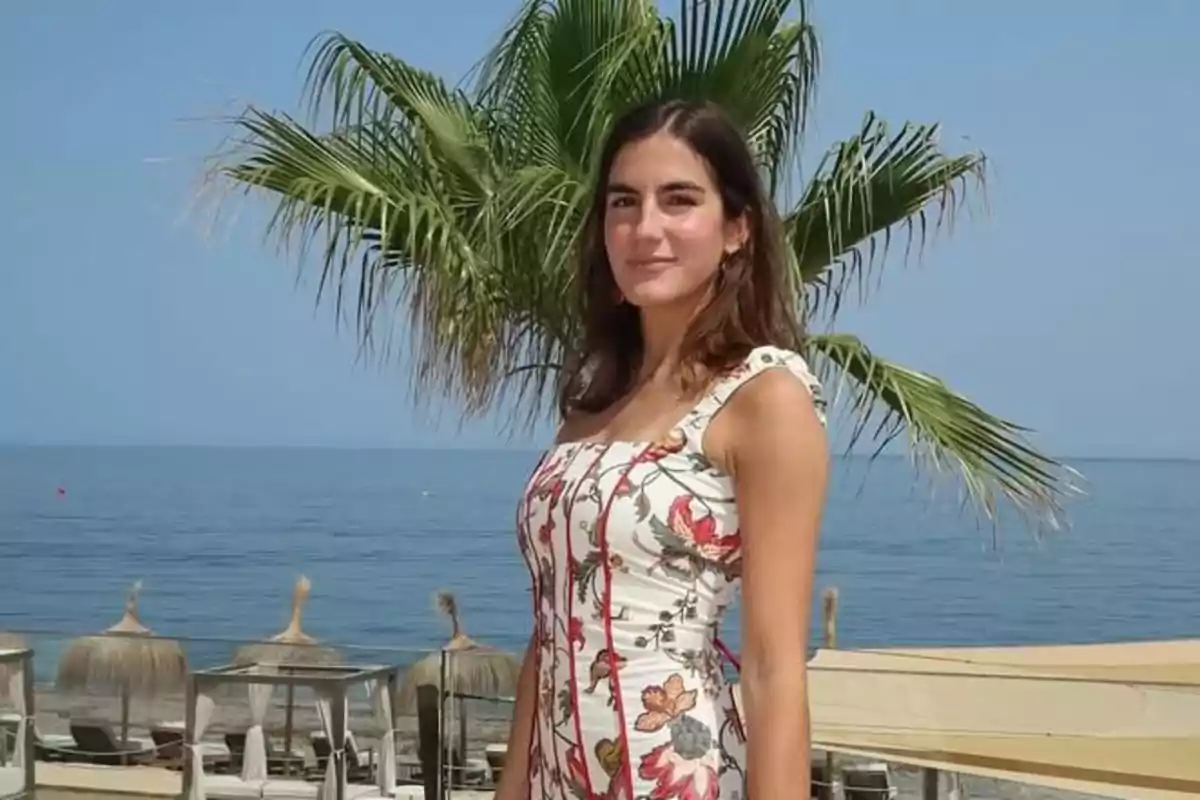 Mujer joven con vestido floral posando frente al mar con palmeras y sombrillas de playa al fondo Mujer joven con vestido floral posando frente al mar con palmeras y sombrillas de playa al fondo