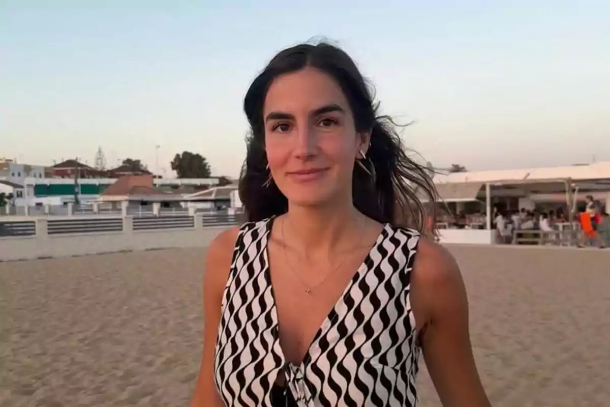 Mujer joven de cabello largo y oscuro sonríe mientras camina por la playa al atardecer con casas y personas al fondo Mujer joven de cabello largo y oscuro sonríe mientras camina por la playa al atardecer con casas y personas al fondo