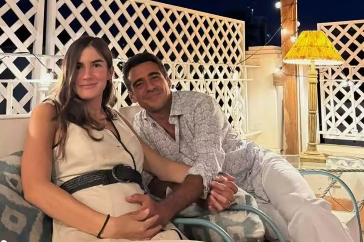 Pareja sentada en una terraza iluminada por una lámpara, ambos sonríen y la mujer sostiene su vientre mientras el hombre le toma la mano