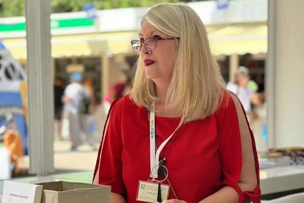 Mujer de cabello rubio y lacio con gafas y blusa roja, de pie junto a una mesa en un entorno luminoso y concurrido Mujer de cabello rubio y lacio con gafas y blusa roja, de pie junto a una mesa en un entorno luminoso y concurrido