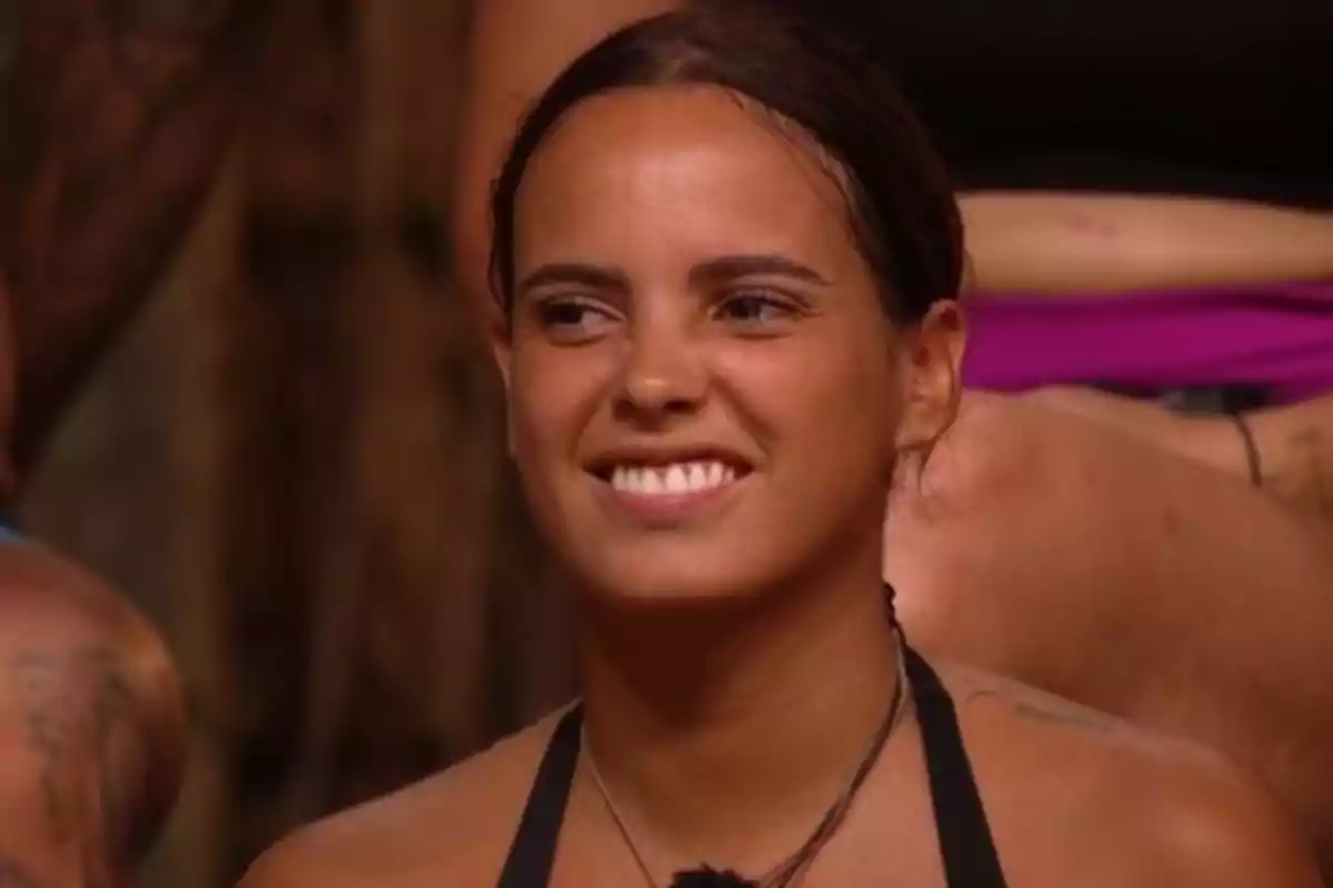 Gloria Camila joven de cabello oscuro recogido sonríe mientras está sentada en un ambiente interior con otras personas alrededor Gloria Camila joven de cabello oscuro recogido sonríe mientras está sentada en un ambiente interior con otras personas alrededor