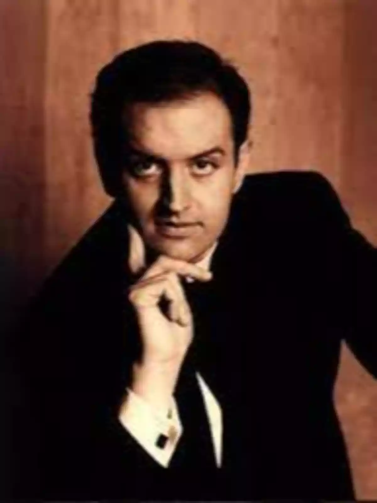 Hombre de cabello oscuro con traje negro posando con la mano en el mentón frente a un fondo marrón Hombre de cabello oscuro con traje negro posando con la mano en el mentón frente a un fondo marrón