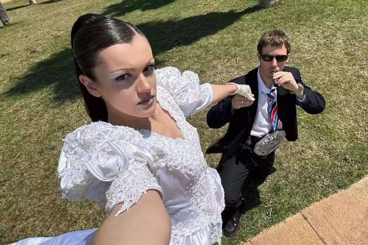 Una mujer con vestido blanco y guantes toma una selfie al aire libre junto a un hombre con traje oscuro, corbata y gafas de sol que sostiene una petaca mientras ambos posan sobre césped bajo el sol