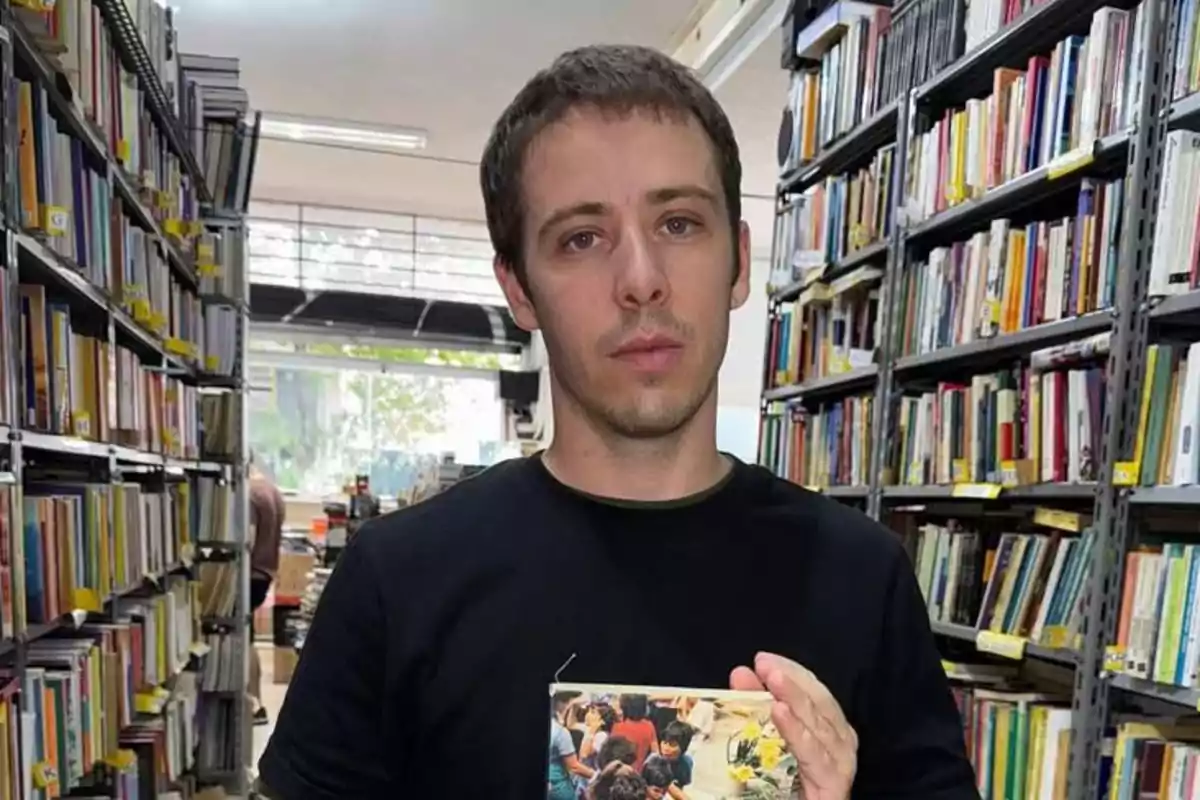 Hombre joven de cabello corto y camiseta oscura sostiene un libro en una biblioteca rodeado de estantes llenos de libros Hombre joven de cabello corto y camiseta oscura sostiene un libro en una biblioteca rodeado de estantes llenos de libros