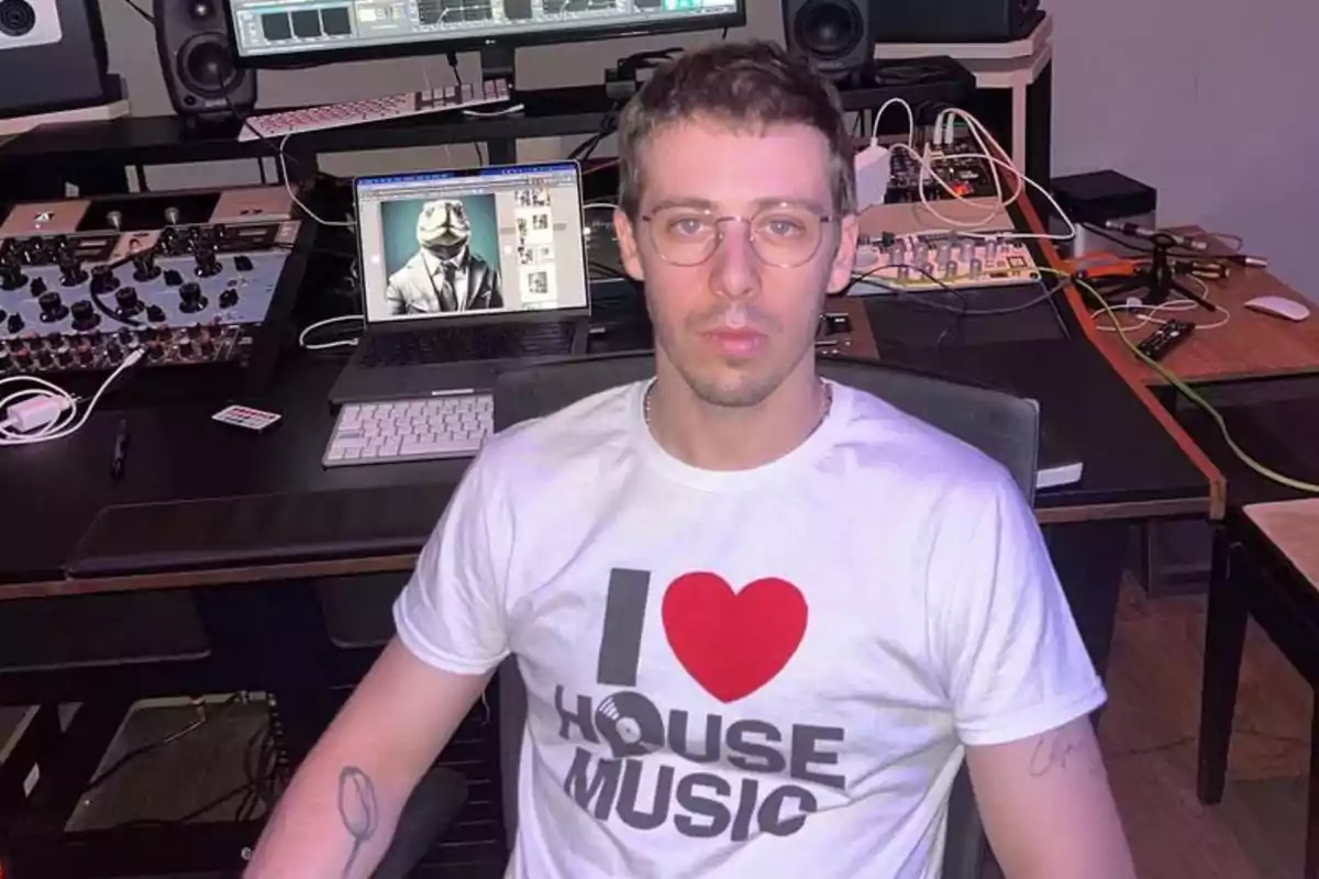 Hombre con gafas y camiseta blanca que dice I love house music sentado en un estudio de música con equipo de audio y una laptop al fondo Hombre con gafas y camiseta blanca que dice I love house music sentado en un estudio de música con equipo de audio y una laptop al fondo