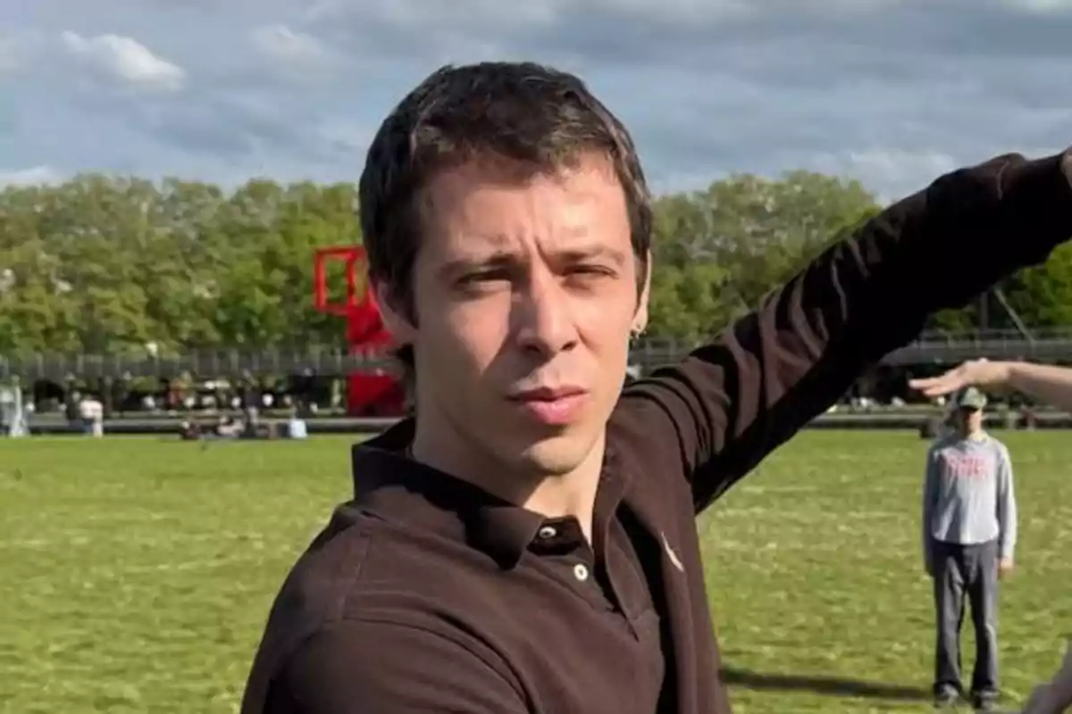 Joven de cabello corto y camisa oscura posando al aire libre en un parque con césped y árboles al fondo Joven de cabello corto y camisa oscura posando al aire libre en un parque con césped y árboles al fondo