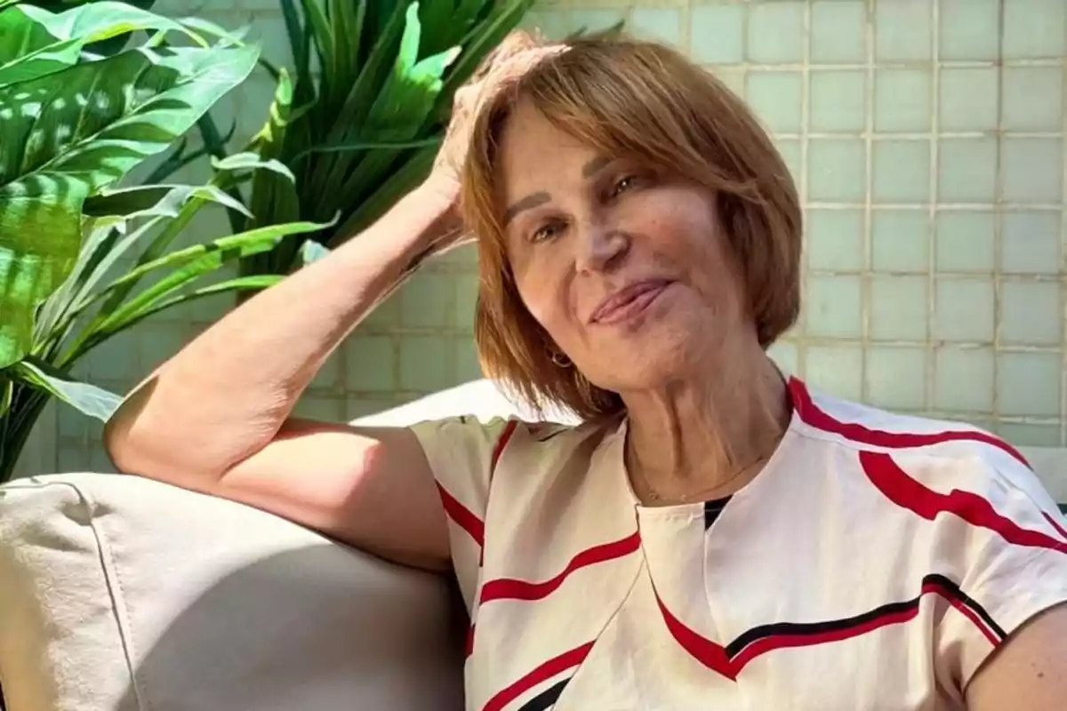 Mujer mayor de cabello corto y castaño sentada en un sillón claro con plantas verdes al fondo y luz natural iluminando su rostro