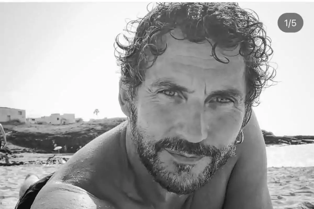 Hombre de cabello rizado y barba recostado en la arena de una playa mirando a la cámara en una imagen en blanco y negro