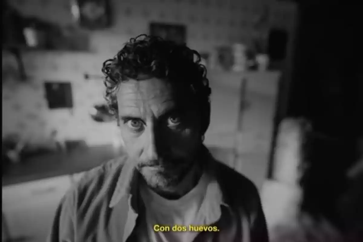 Hombre de cabello rizado y barba mirando fijamente a la cámara en una cocina con la frase Con dos huevos escrita en la parte inferior Hombre de cabello rizado y barba mirando fijamente a la cámara en una cocina con la frase Con dos huevos escrita en la parte inferior