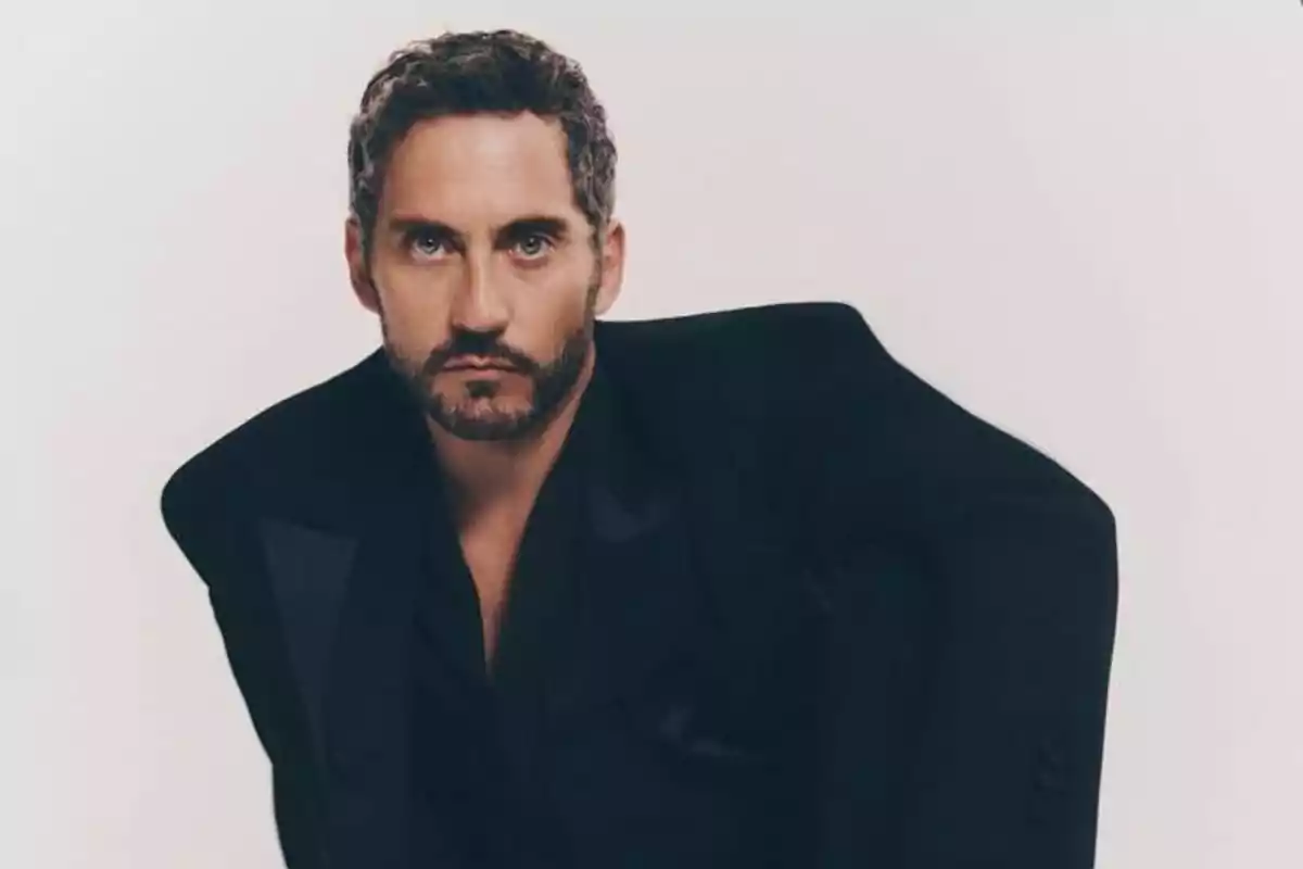 Hombre de cabello corto y barba con saco negro posando frente a un fondo claro Hombre de cabello corto y barba con saco negro posando frente a un fondo claro