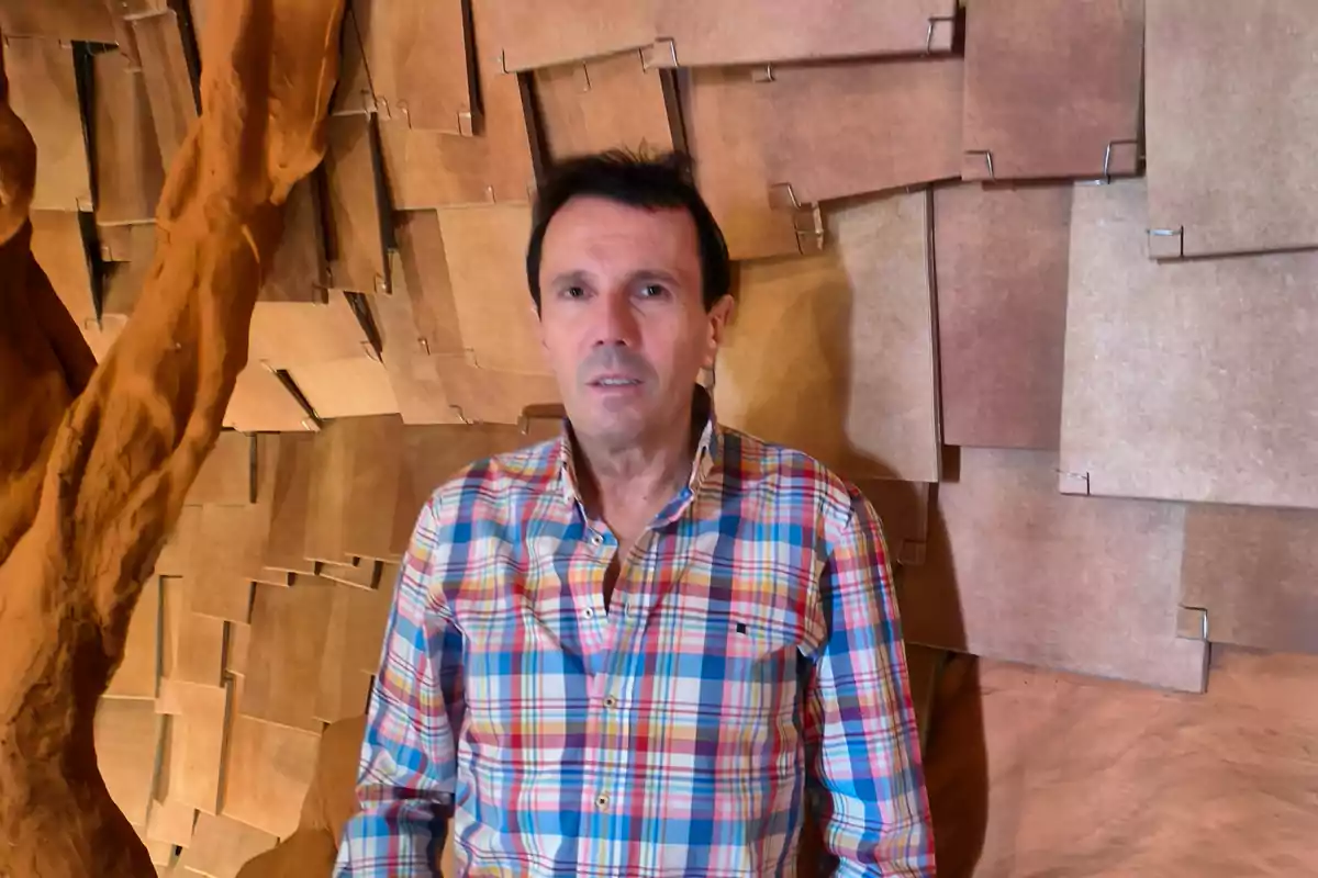 Hombre con camisa de cuadros coloridos posando frente a una pared de paneles marrones con textura geométrica