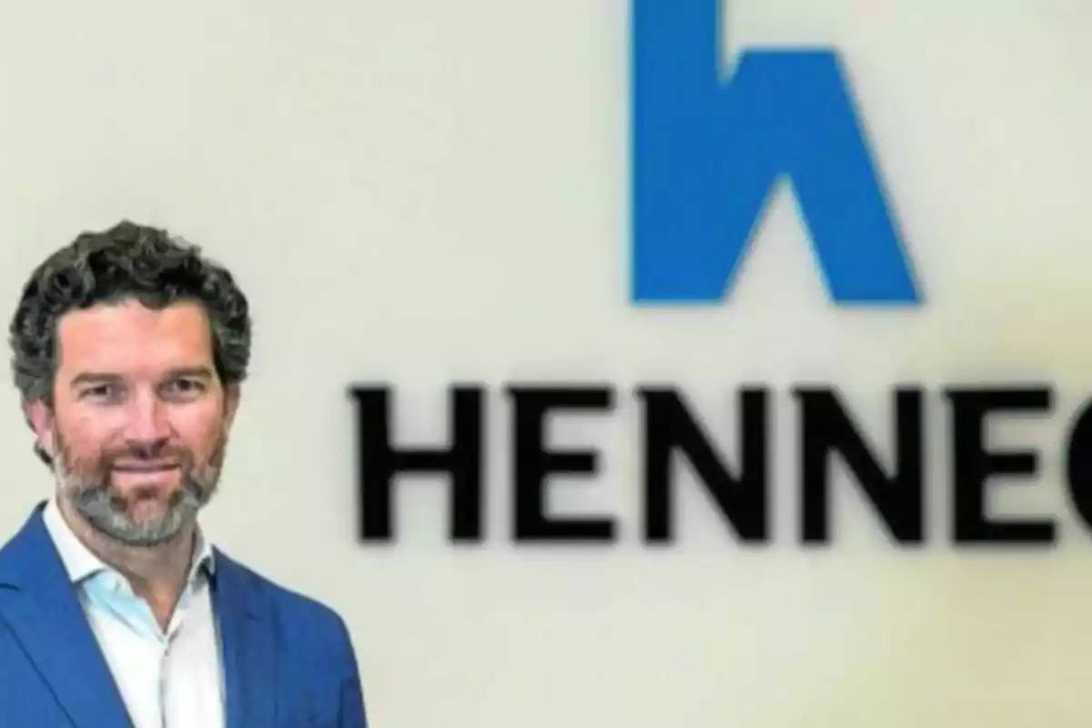 Hombre de cabello rizado y barba con saco azul posando frente a un logotipo azul y negro en una pared blanca que dice HENNEC Hombre de cabello rizado y barba con saco azul posando frente a un logotipo azul y negro en una pared blanca que dice HENNEC