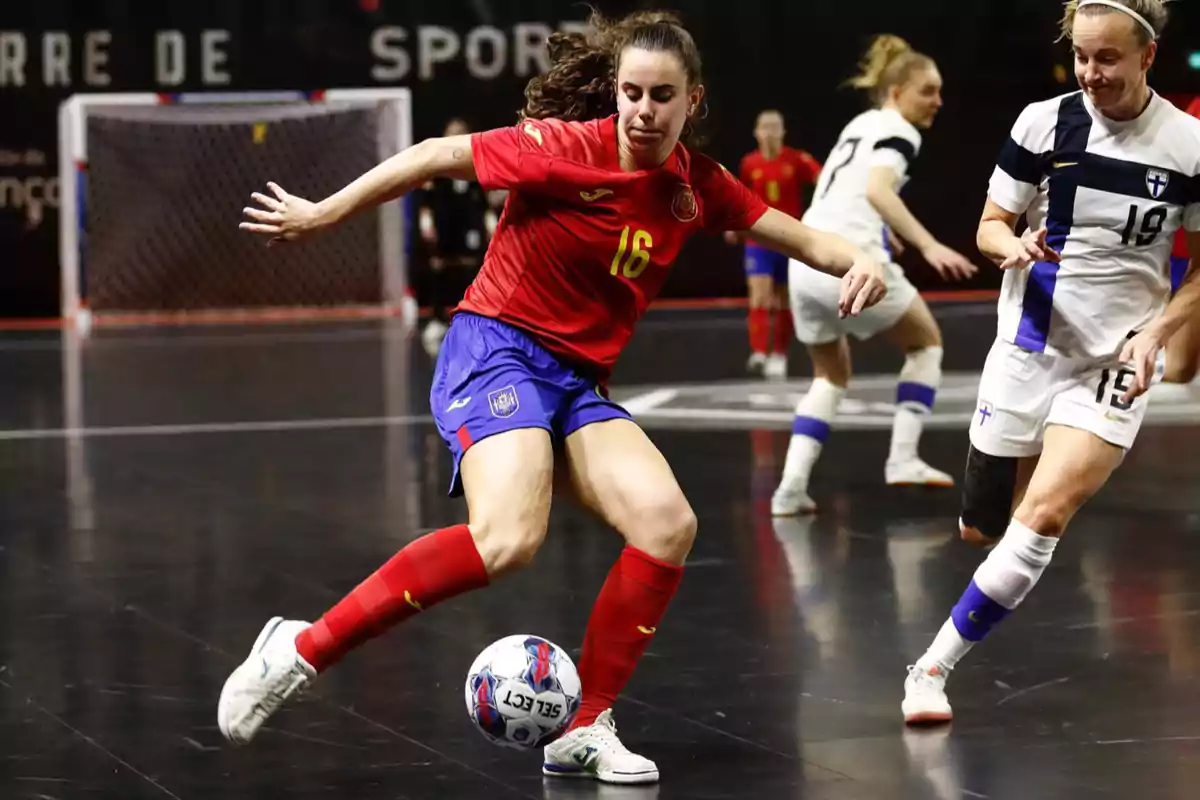 Jugadora de fútbol sala con uniforme rojo y azul controla el balón mientras una rival con uniforme blanco la presiona en una cancha cubierta Jugadora de fútbol sala con uniforme rojo y azul controla el balón mientras una rival con uniforme blanco la presiona en una cancha cubierta