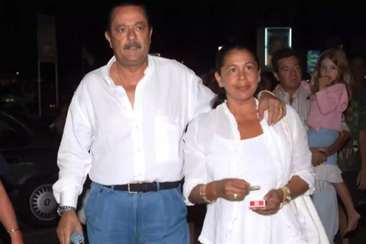 Una pareja vestida de blanco camina junta de noche mientras un hombre sostiene a una niña en el fondo Una pareja vestida de blanco camina junta de noche mientras un hombre sostiene a una niña en el fondo