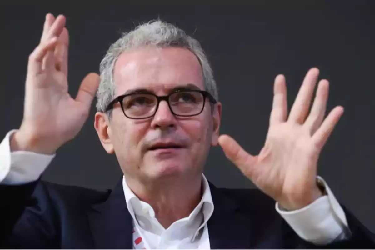 Hombre de cabello canoso y gafas levantando ambas manos mientras habla, vistiendo saco oscuro y camisa blanca, fondo oscuro. Hombre de cabello canoso y gafas levantando ambas manos mientras habla, vistiendo saco oscuro y camisa blanca, fondo oscuro.