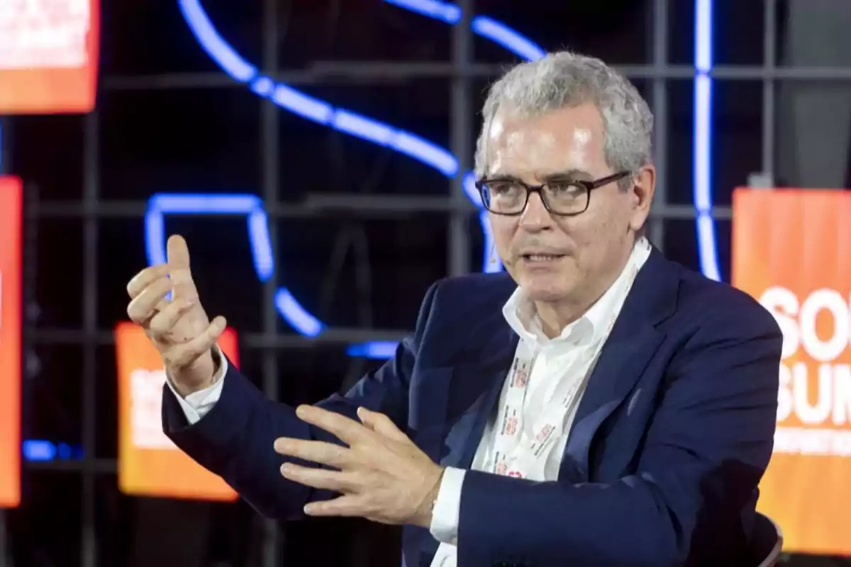 Hombre de cabello canoso y gafas hablando y gesticulando con las manos en un evento con fondo de luces y carteles naranjas Hombre de cabello canoso y gafas hablando y gesticulando con las manos en un evento con fondo de luces y carteles naranjas