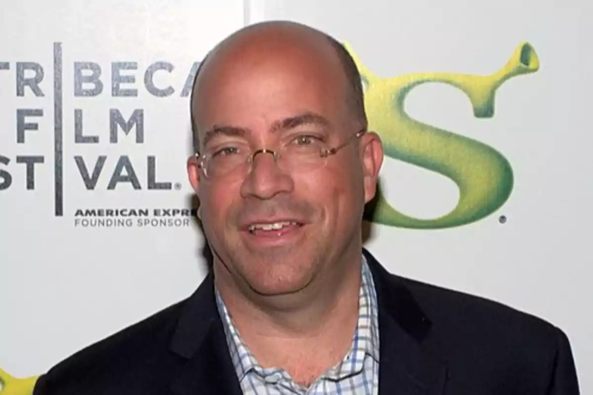 Hombre calvo con gafas y saco oscuro sonriendo frente a un fondo con logotipos de un festival de cine y la película Shrek Hombre calvo con gafas y saco oscuro sonriendo frente a un fondo con logotipos de un festival de cine y la película Shrek