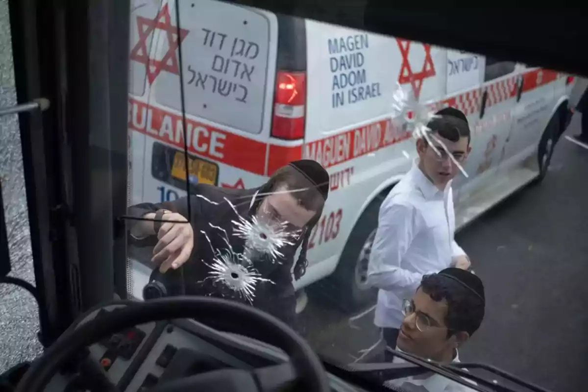 Tres jóvenes observan los impactos de bala en el parabrisas de un vehículo mientras una ambulancia israelí está estacionada al fondo Tres jóvenes observan los impactos de bala en el parabrisas de un vehículo mientras una ambulancia israelí está estacionada al fondo