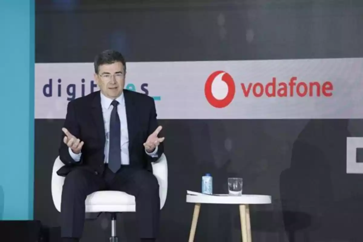 Hombre con traje sentado en una silla blanca durante un evento, con logotipos de empresas en el fondo y una mesa pequeña con una lata y un vaso de agua a su lado Hombre con traje sentado en una silla blanca durante un evento, con logotipos de empresas en el fondo y una mesa pequeña con una lata y un vaso de agua a su lado