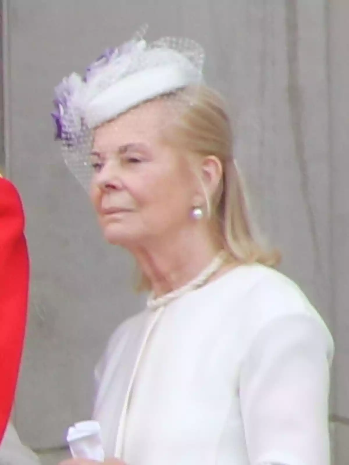 Mujer mayor con sombrero blanco decorado con velo y flores, vestida elegantemente con un conjunto claro y collar de perlas, de pie frente a una pared gris Mujer mayor con sombrero blanco decorado con velo y flores, vestida elegantemente con un conjunto claro y collar de perlas, de pie frente a una pared gris