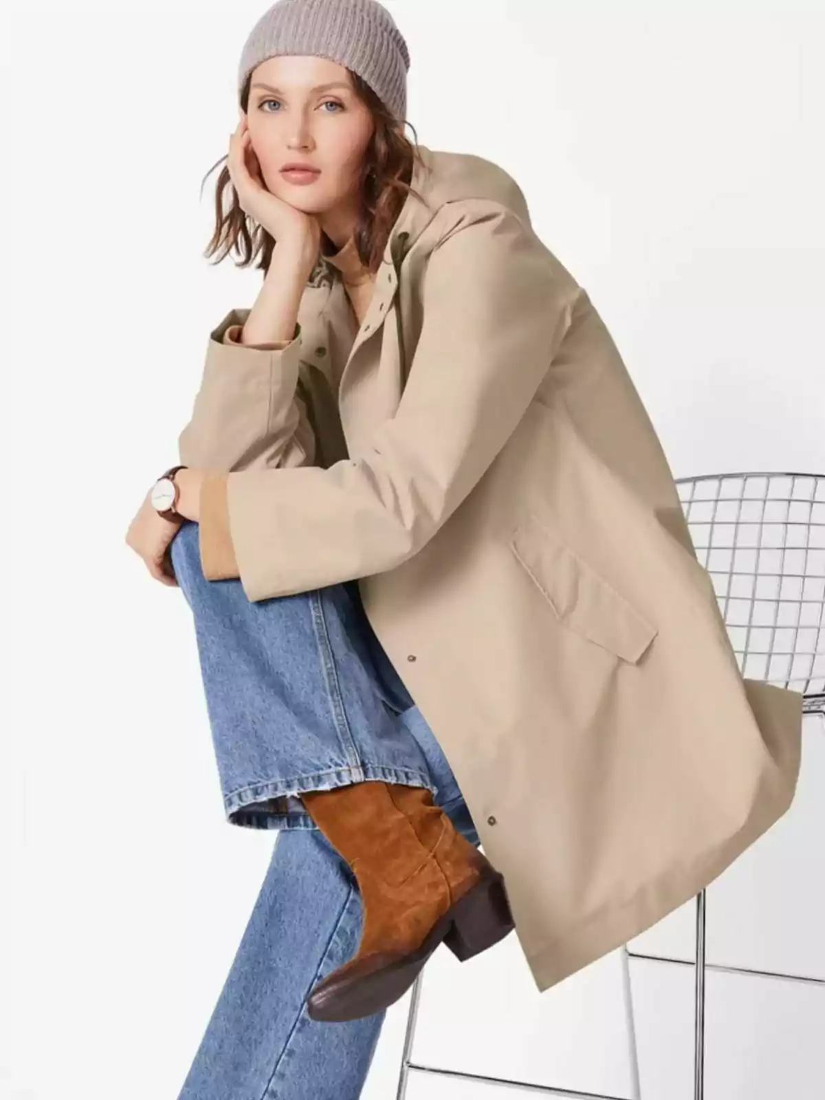 Mujer sentada en una silla de metal, lleva un abrigo beige, jeans, botas marrones y un gorro gris, con expresión pensativa y la cabeza apoyada en una mano. Mujer sentada en una silla de metal, lleva un abrigo beige, jeans, botas marrones y un gorro gris, con expresión pensativa y la cabeza apoyada en una mano.