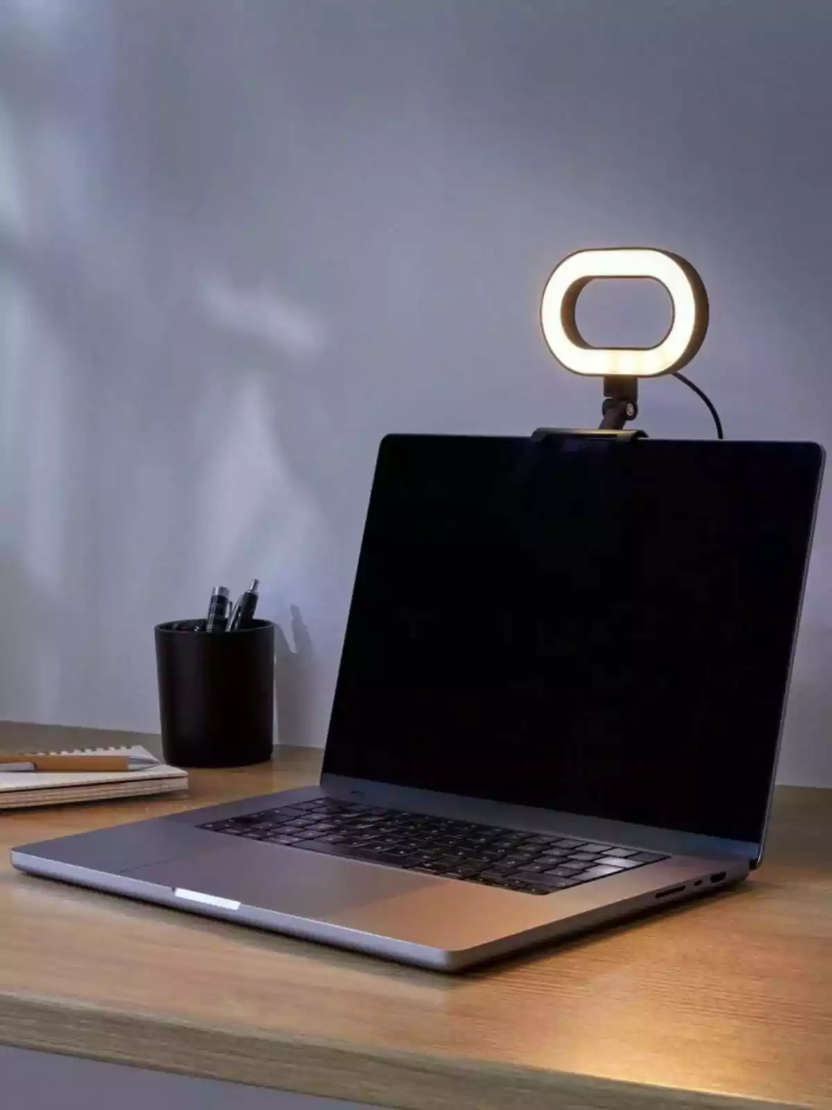 Laptop sobre un escritorio de madera con una luz de aro encendida colocada en la parte superior de la pantalla y un vaso con bolígrafos al fondo Laptop sobre un escritorio de madera con una luz de aro encendida colocada en la parte superior de la pantalla y un vaso con bolígrafos al fondo