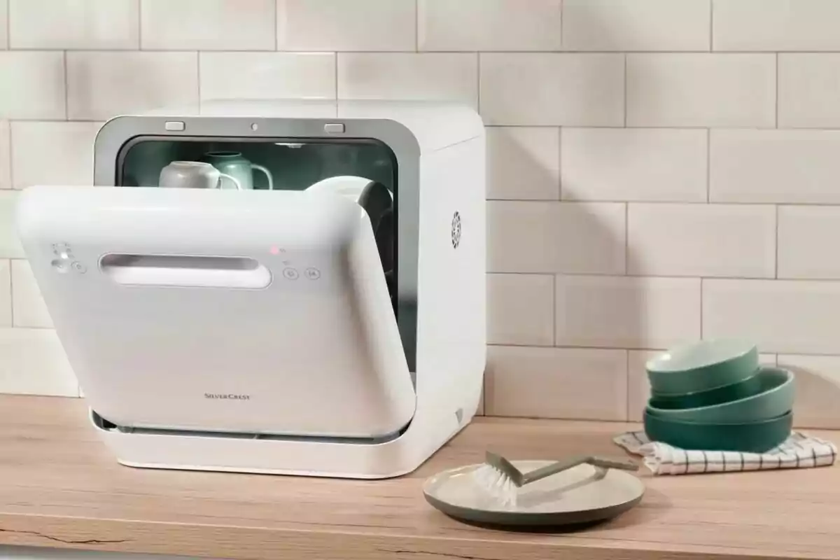 Lavavajillas compacto de color blanco sobre una encimera de madera con la puerta abierta y algunos platos y tazas en su interior, junto a un plato, un cepillo y varios tazones apilados a un lado Lavavajillas compacto de color blanco sobre una encimera de madera con la puerta abierta y algunos platos y tazas en su interior, junto a un plato, un cepillo y varios tazones apilados a un lado