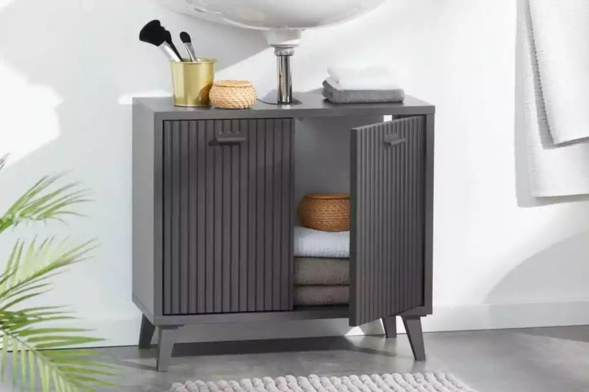 Mueble gris de baño con puertas abiertas, toallas dobladas y cestas organizadoras, junto a un lavabo y accesorios de tocador sobre la superficie Mueble gris de baño con puertas abiertas, toallas dobladas y cestas organizadoras, junto a un lavabo y accesorios de tocador sobre la superficie