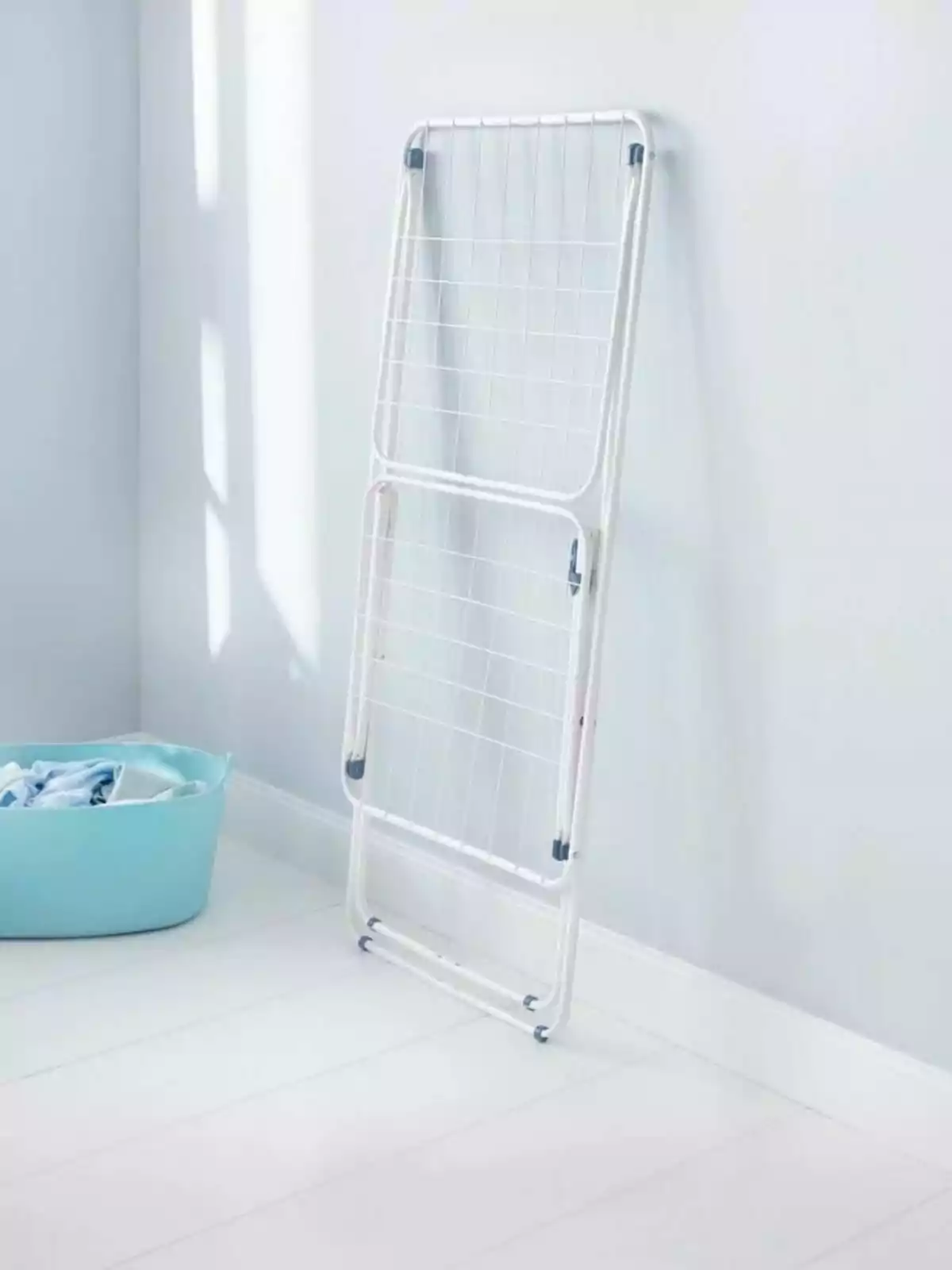 Tendedero plegable de metal blanco apoyado en la pared junto a una cesta azul con ropa en una habitación iluminada Tendedero plegable de metal blanco apoyado en la pared junto a una cesta azul con ropa en una habitación iluminada