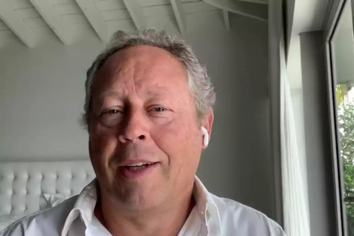 Hombre de cabello canoso usando auriculares inalámbricos y camisa blanca en una habitación iluminada con ventana grande Hombre de cabello canoso usando auriculares inalámbricos y camisa blanca en una habitación iluminada con ventana grande