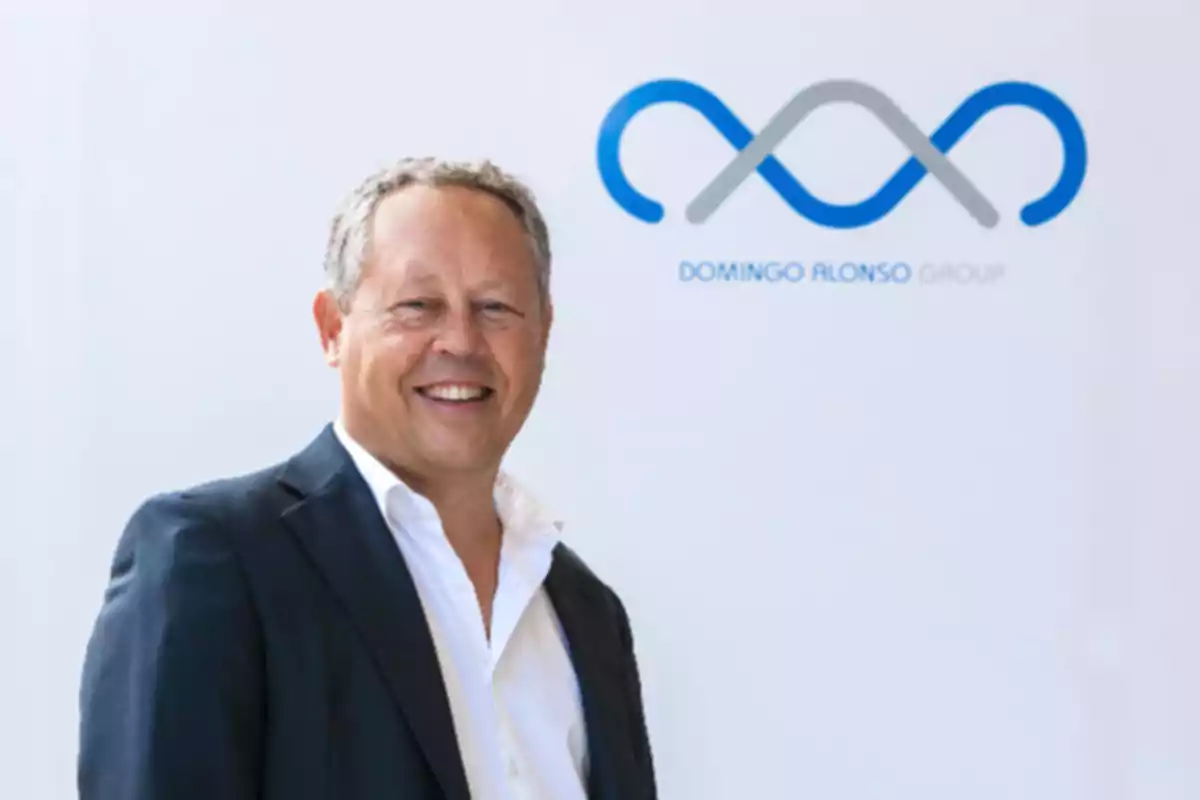 Hombre de traje sonriente posando frente a un logotipo de Domingo Alonso Group sobre fondo claro