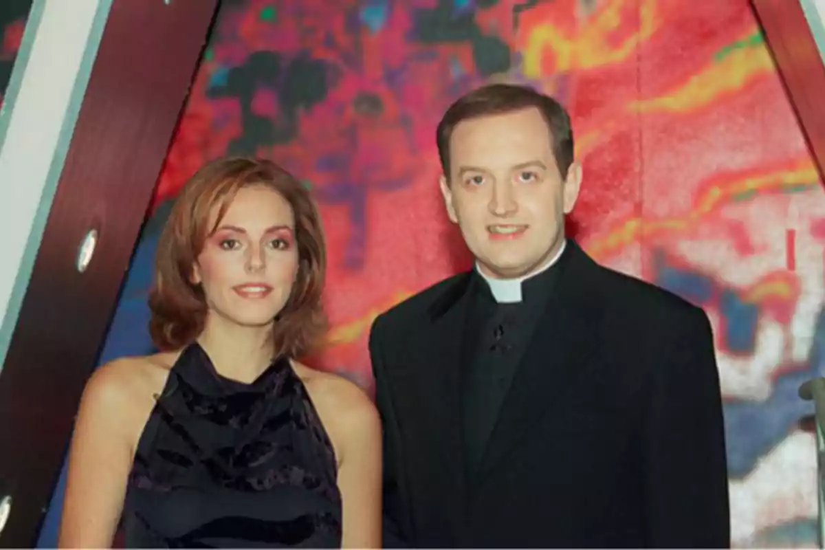 Una mujer con vestido negro y un hombre vestido de sacerdote posan juntos frente a un fondo colorido Una mujer con vestido negro y un hombre vestido de sacerdote posan juntos frente a un fondo colorido