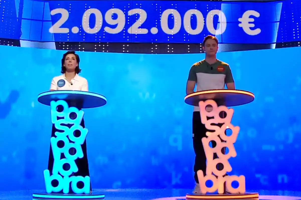 Rosa y Manu de pie en sus atriles iluminados en un plató azul de un concurso de televisión, con un premio de más de dos millones de euros mostrado en la pantalla superior. Captura de 'Pasapalabra' Rosa y Manu de pie en sus atriles iluminados en un plató azul de un concurso de televisión, con un premio de más de dos millones de euros mostrado en la pantalla superior. Captura de 'Pasapalabra'