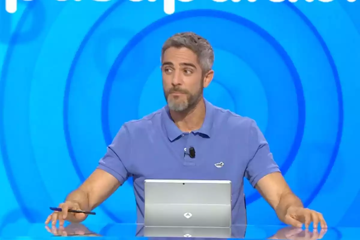 Roberto Leal sentado frente a una mesa con una tablet y un bolígrafo en la mano, fondo azul con círculos concéntricos. Captura de 'Pasapalabra' Roberto Leal sentado frente a una mesa con una tablet y un bolígrafo en la mano, fondo azul con círculos concéntricos. Captura de 'Pasapalabra'