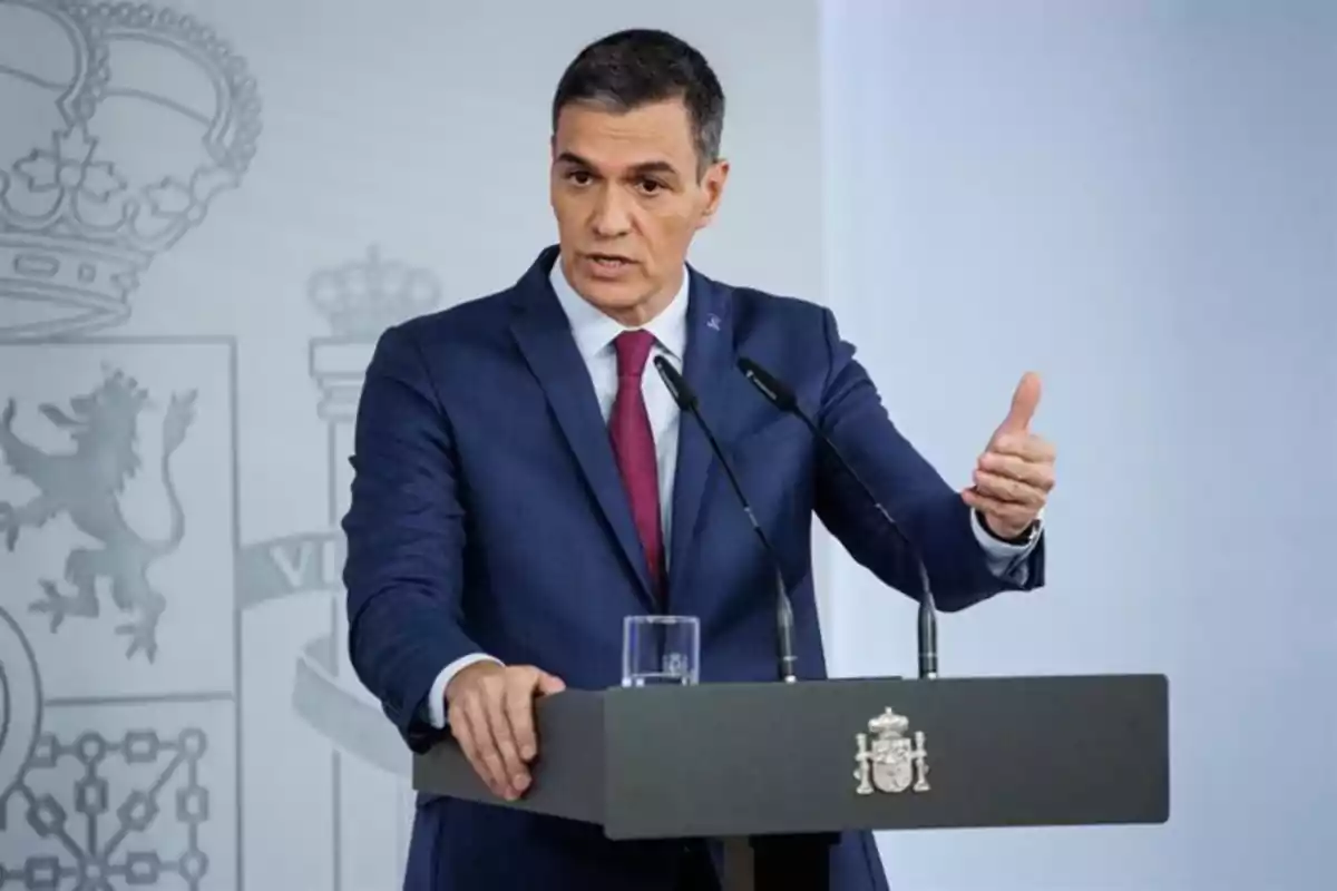 Hombre con traje azul y corbata roja hablando en una rueda de prensa detrás de un atril con el escudo de España