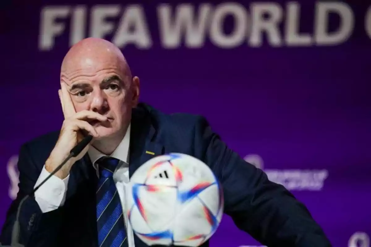 Hombre calvo con traje oscuro sentado frente a un micrófono y un balón de fútbol, con fondo morado y letras borrosas de FIFA World Cup. Hombre calvo con traje oscuro sentado frente a un micrófono y un balón de fútbol, con fondo morado y letras borrosas de FIFA World Cup.