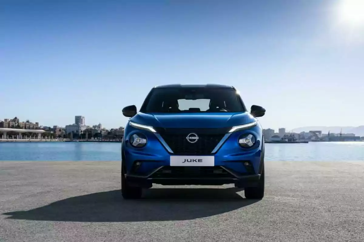 Auto Nissan Juke azul visto de frente estacionado junto al mar con una ciudad al fondo bajo un cielo despejado Auto Nissan Juke azul visto de frente estacionado junto al mar con una ciudad al fondo bajo un cielo despejado