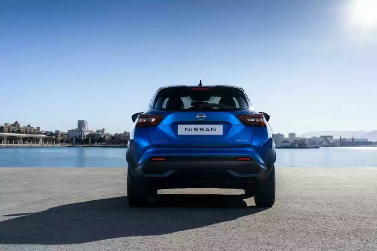 Auto Nissan azul visto desde atrás estacionado frente al mar con una ciudad al fondo y cielo despejado Auto Nissan azul visto desde atrás estacionado frente al mar con una ciudad al fondo y cielo despejado
