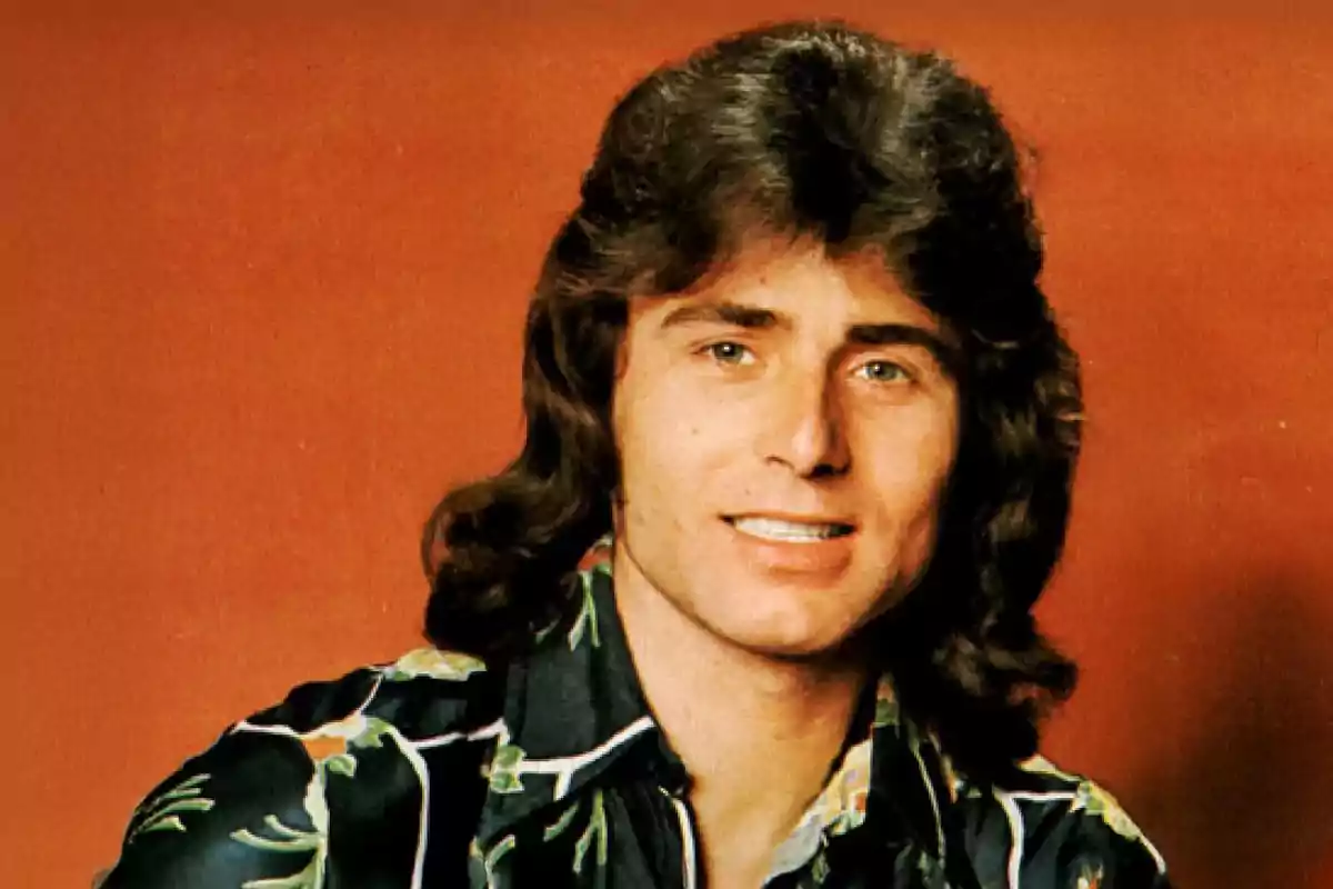 Hombre joven de cabello largo y oscuro con camisa estampada posando frente a un fondo naranja Hombre joven de cabello largo y oscuro con camisa estampada posando frente a un fondo naranja