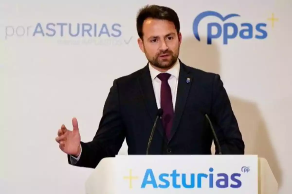 Hombre con barba y traje oscuro hablando en un atril con el logo de Asturias y el Partido Popular de Asturias al fondo