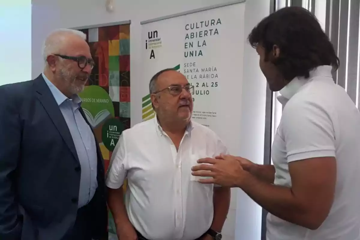 Tres hombres conversan en un evento académico frente a carteles de la Universidad Internacional de Andalucía Tres hombres conversan en un evento académico frente a carteles de la Universidad Internacional de Andalucía