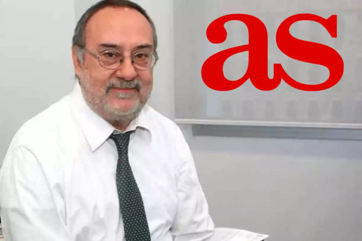 Hombre de barba y gafas con camisa blanca y corbata oscura sentado frente a un fondo con el logo rojo de AS Hombre de barba y gafas con camisa blanca y corbata oscura sentado frente a un fondo con el logo rojo de AS