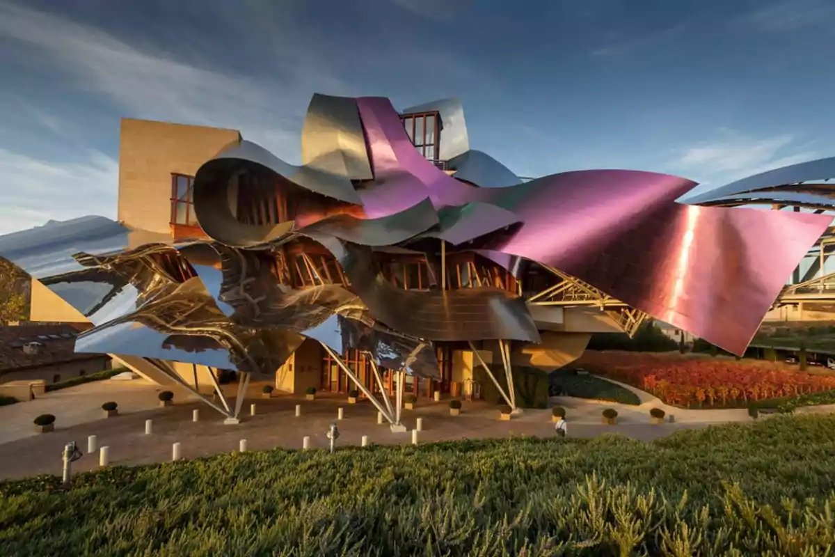 Edificio moderno con formas onduladas y paneles metálicos de colores brillantes rodeado de vegetación y bajo un cielo azul Edificio moderno con formas onduladas y paneles metálicos de colores brillantes rodeado de vegetación y bajo un cielo azul