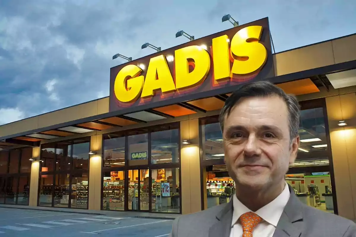 Un hombre con traje y corbata está frente a la entrada de un supermercado Gadis al atardecer